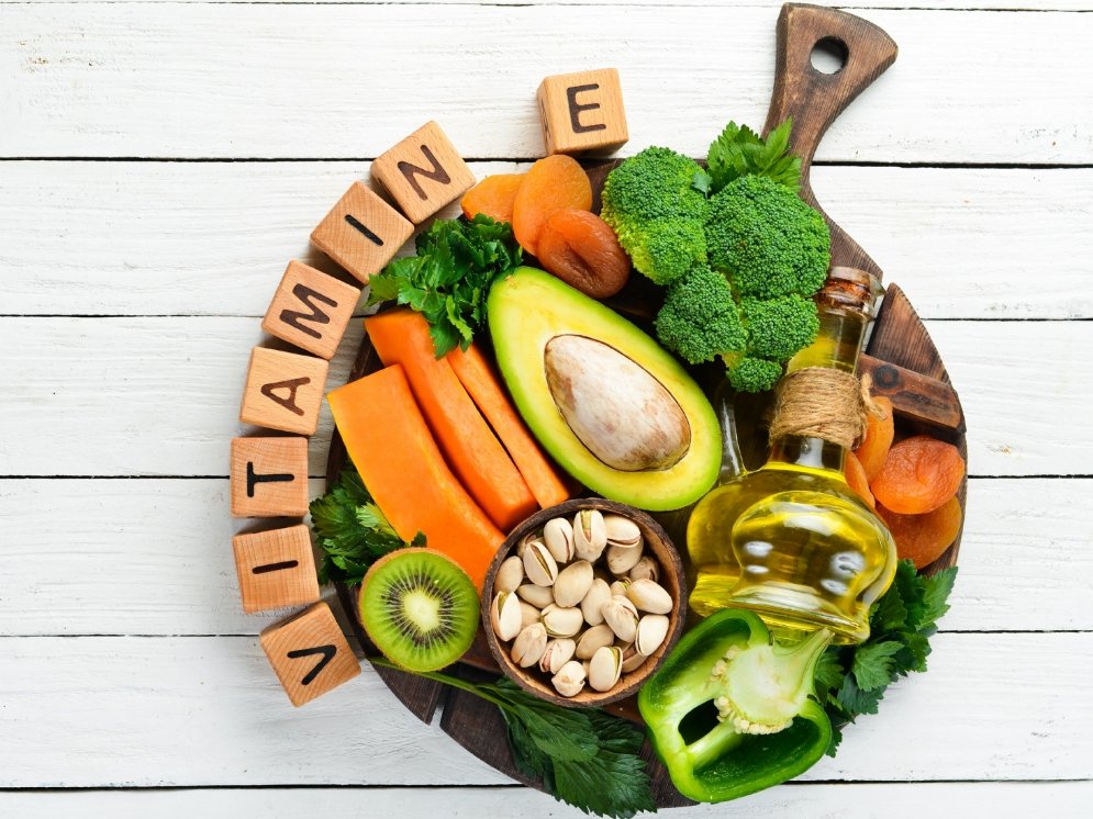 Holzbrettchen mit verschiedenen pflanzlichen Lebensmitteln, wie Avocado, Brokkoli, Pistazien und Paprika, die reich an Vitamin E sind.
