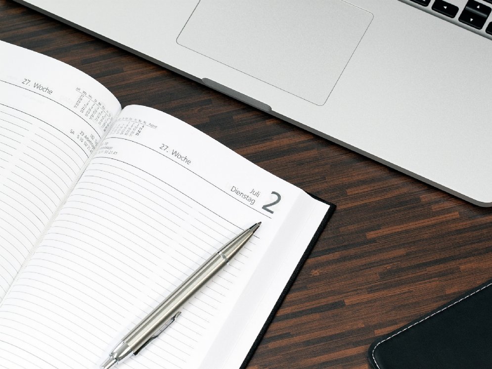 Ein geöffneter Laptop liegt zusammen mit einem aufgeschlagenen Terminplaner und einem silbernen Kugelschreiber auf einem Schreibtisch.
