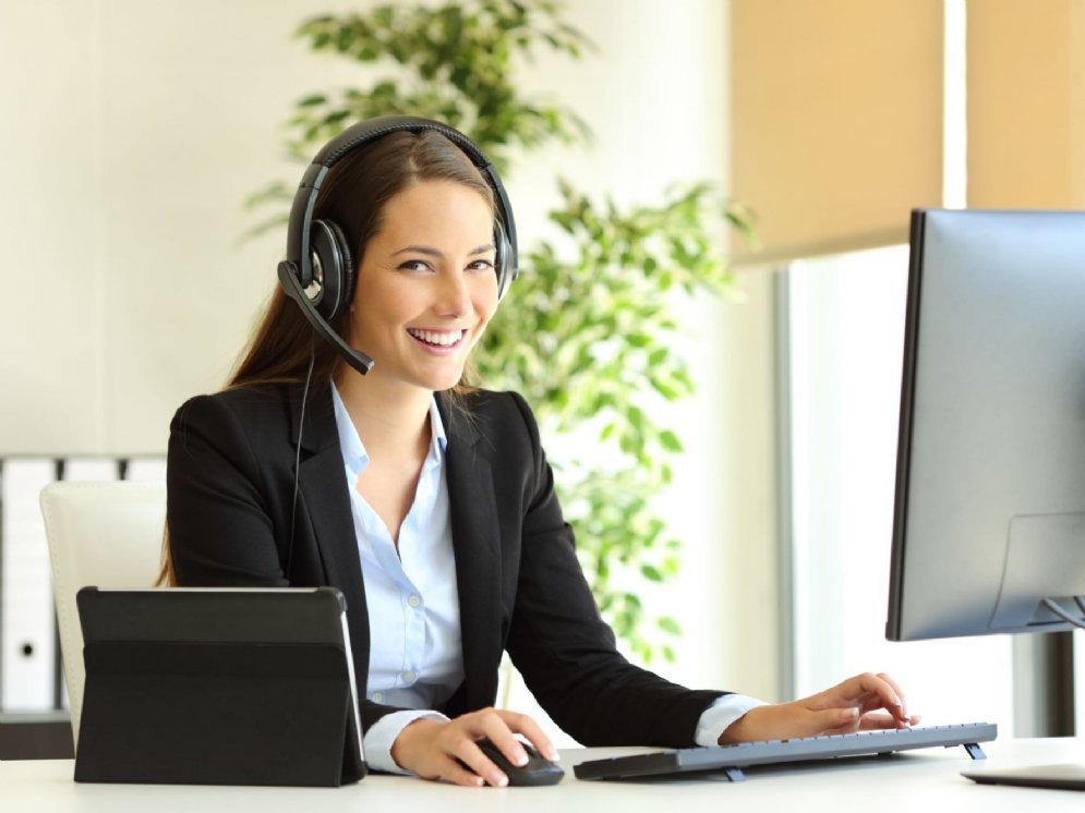 Junge, lächelnde Frau mit Headset sitzt an einem Büroarbeitsplatz.
